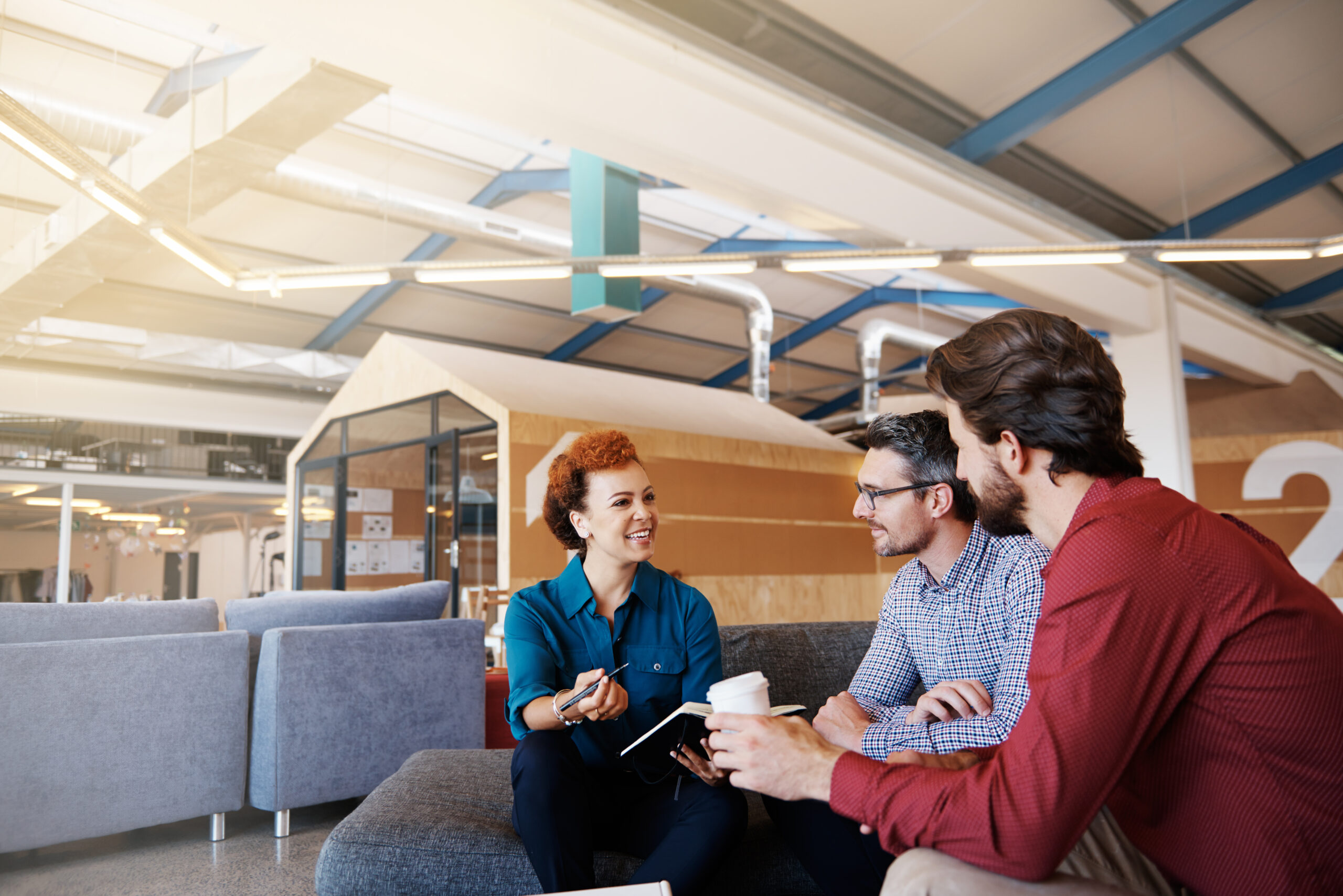 Three people sit on a couch in a modern law office, engaged in conversation. One woman holds a notebook, while two men listen, one holding a coffee cup. The bright workspace features large windows and industrial décor.