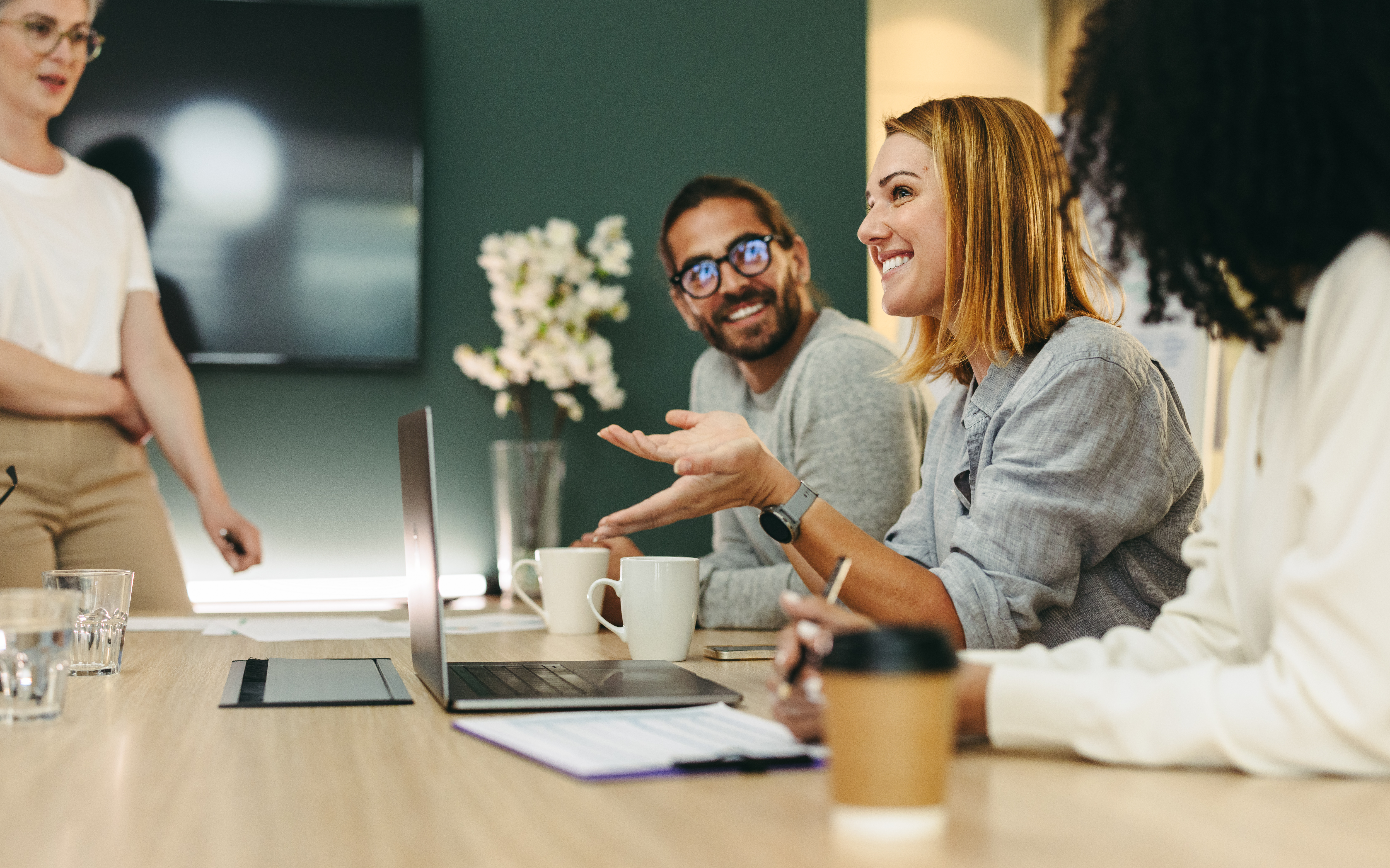 Four people sit around a conference table with laptops, notebooks, and coffee cups, engaged in lively planning discussions. One woman gestures while speaking as the others listen attentively and smile.