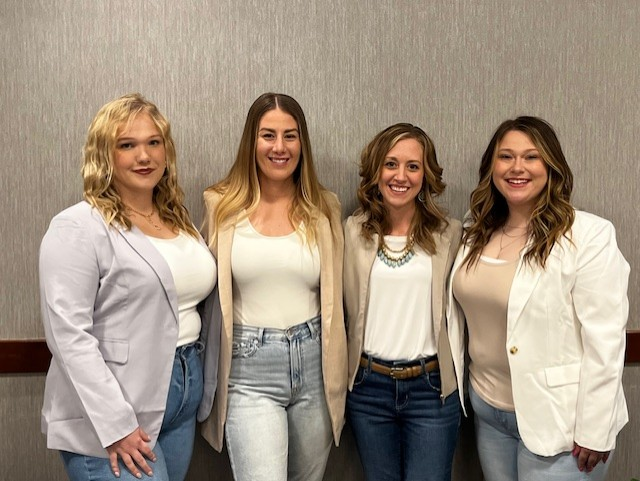 Four women stand side by side in front of a plain wall, smiling at the camera. Dressed in light business blazers and jeans with neutral tops, they have their hands casually by their sides or in their pockets.