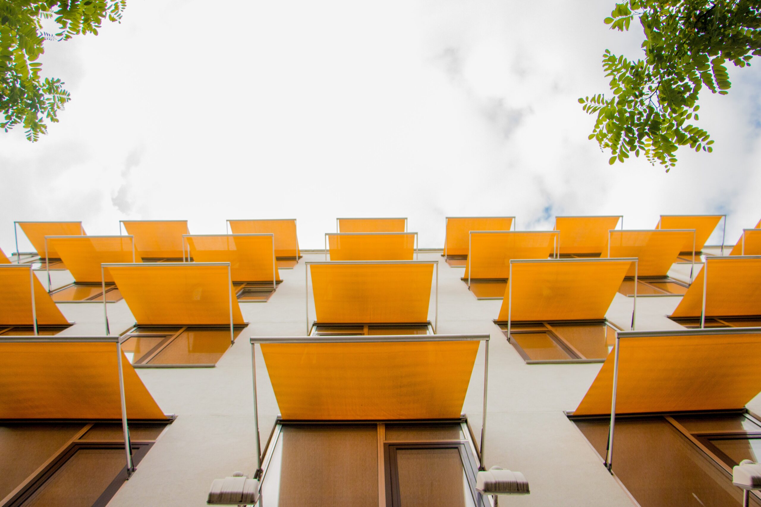 A low-angle view of a modern building with bold orange awnings, set against a partly cloudy sky and framed by green leaves, creates an atmosphere of security—far removed from the world of fraud.