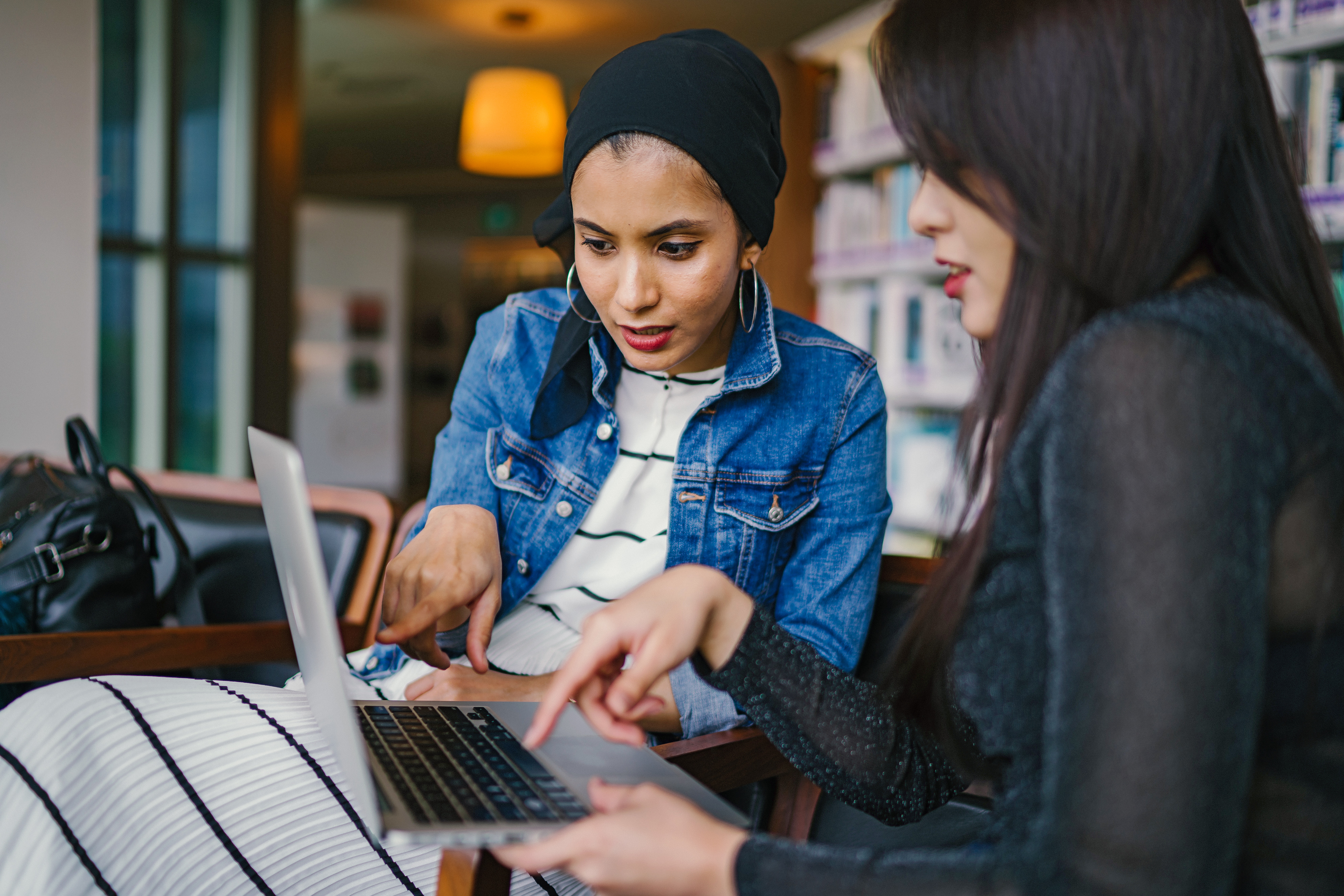 Two women sitting indoors, focused on a laptop. Engaged in a social discussion, one wears a striped dress, denim jacket, and headscarf; the other is dressed in black. Both point at the laptop screen, collaborating closely.