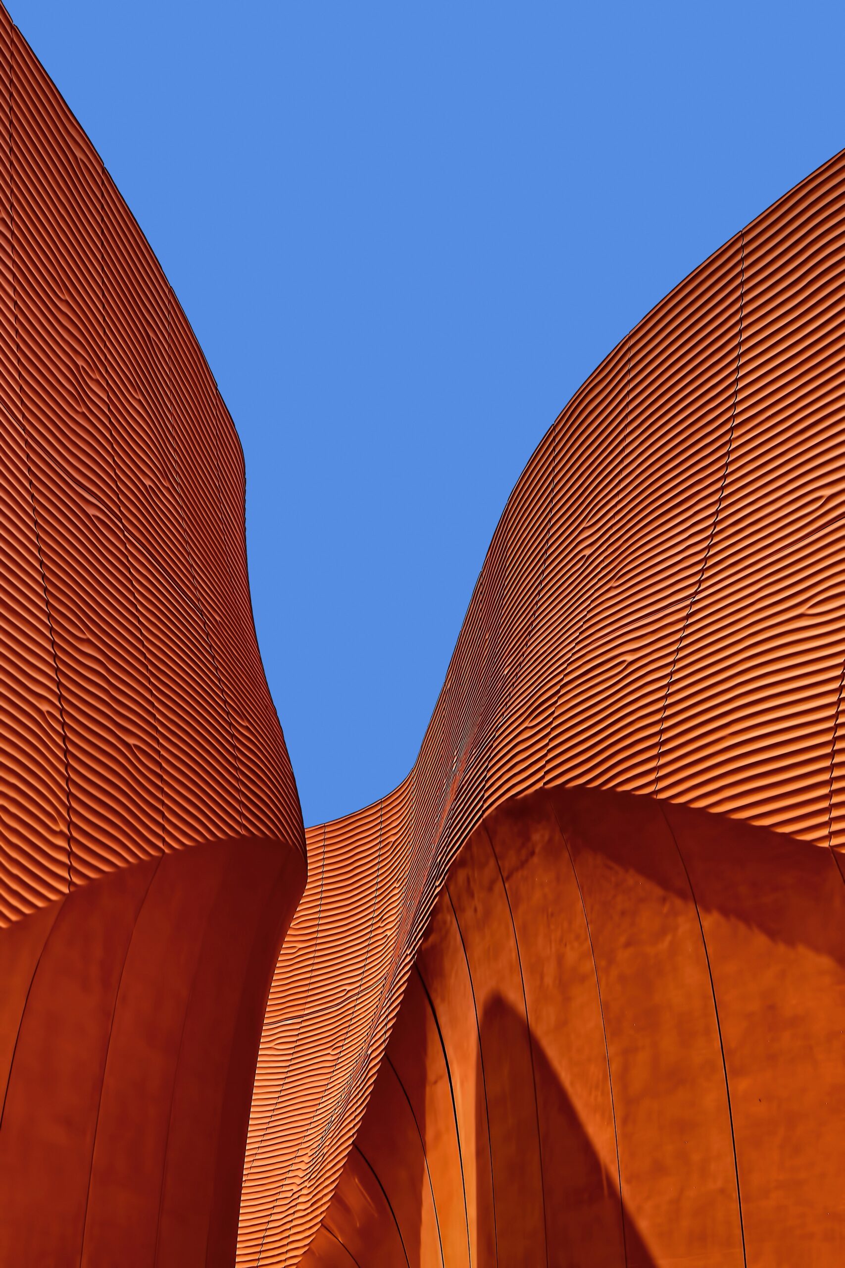 Curved, orange architectural structures with ribbed surfaces rise against a clear blue sky, their dynamic shadows and abstract shapes reflecting the creative spirit often found in self employment.