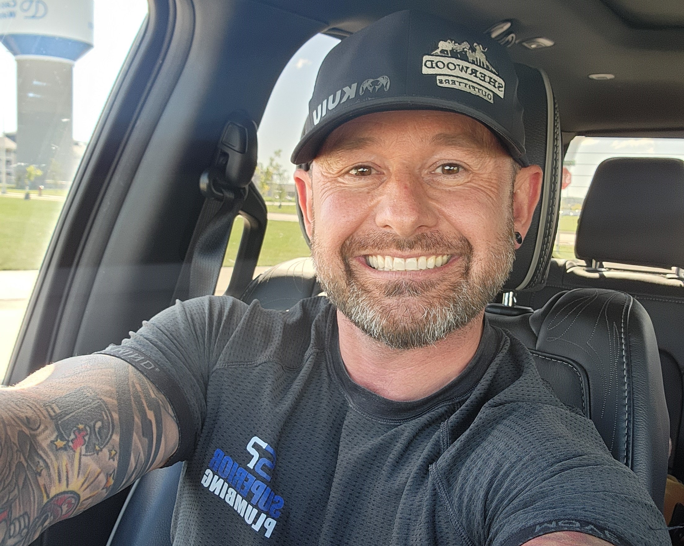 A smiling man with a beard and tattoos takes a selfie in a car. He sports a black shirt that says Superior Plumbing, perfect for plumbing pros, and a black cap with SHERWOOD on it. Sunlight streams through the window.