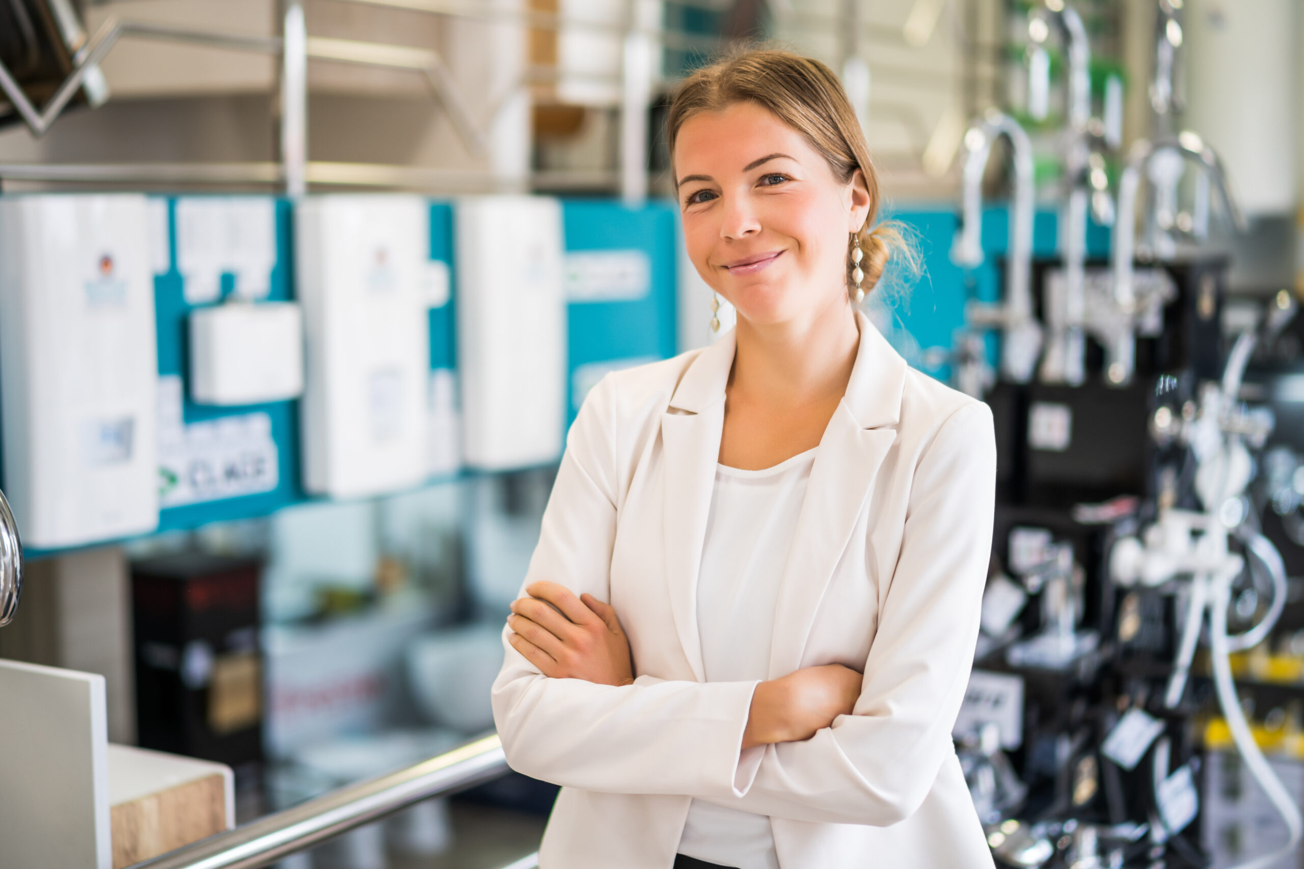 A woman in a white blazer stands with arms crossed, smiling confidently in a modern IT showroom with various appliances and fixtures displayed in the background.