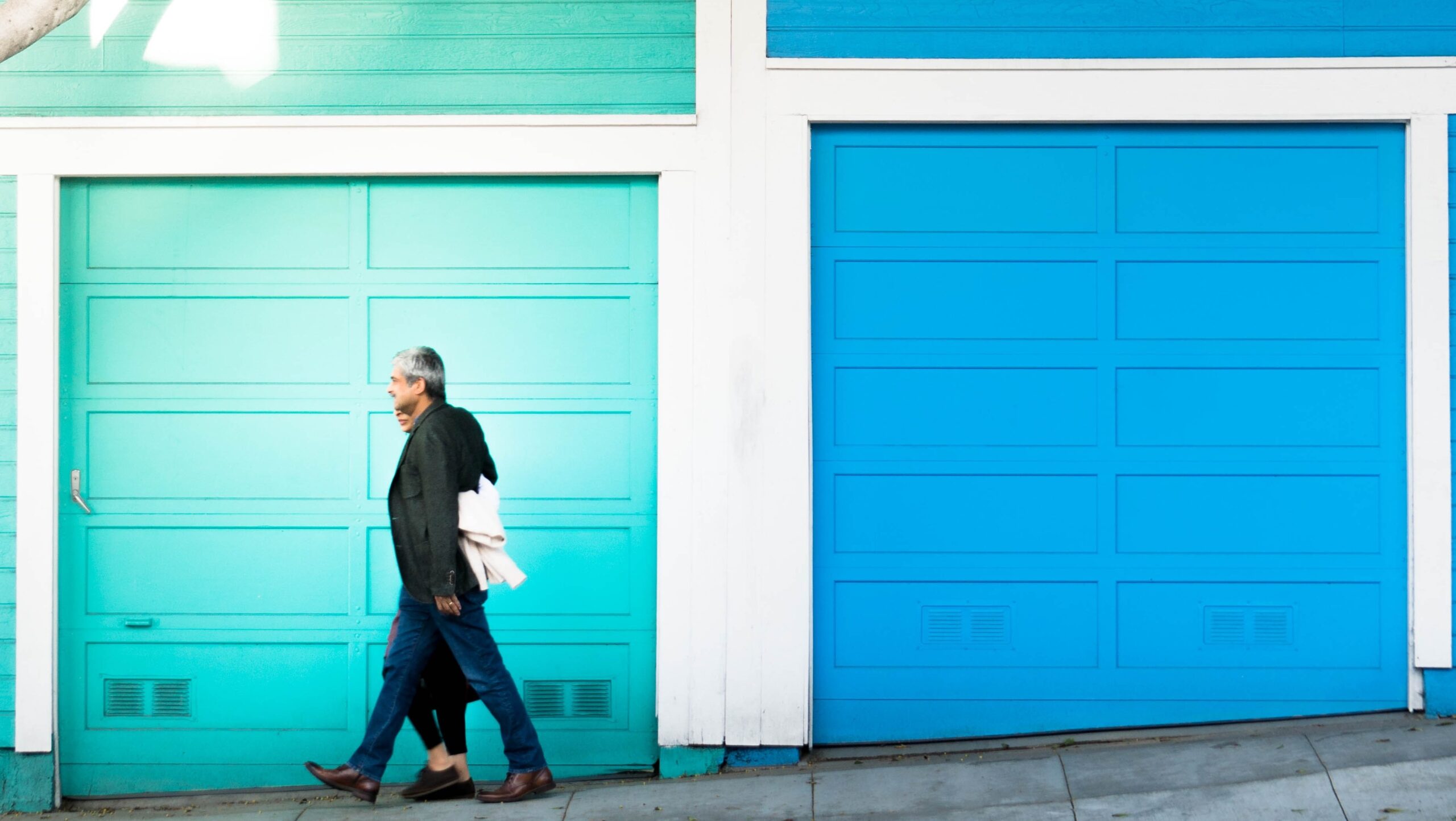 An older man and a younger woman walk side by side past two brightly colored garage doors, one teal and one blue, on a sunny day. The slightly sloped ground invites deductions about the lively neighborhood’s character.