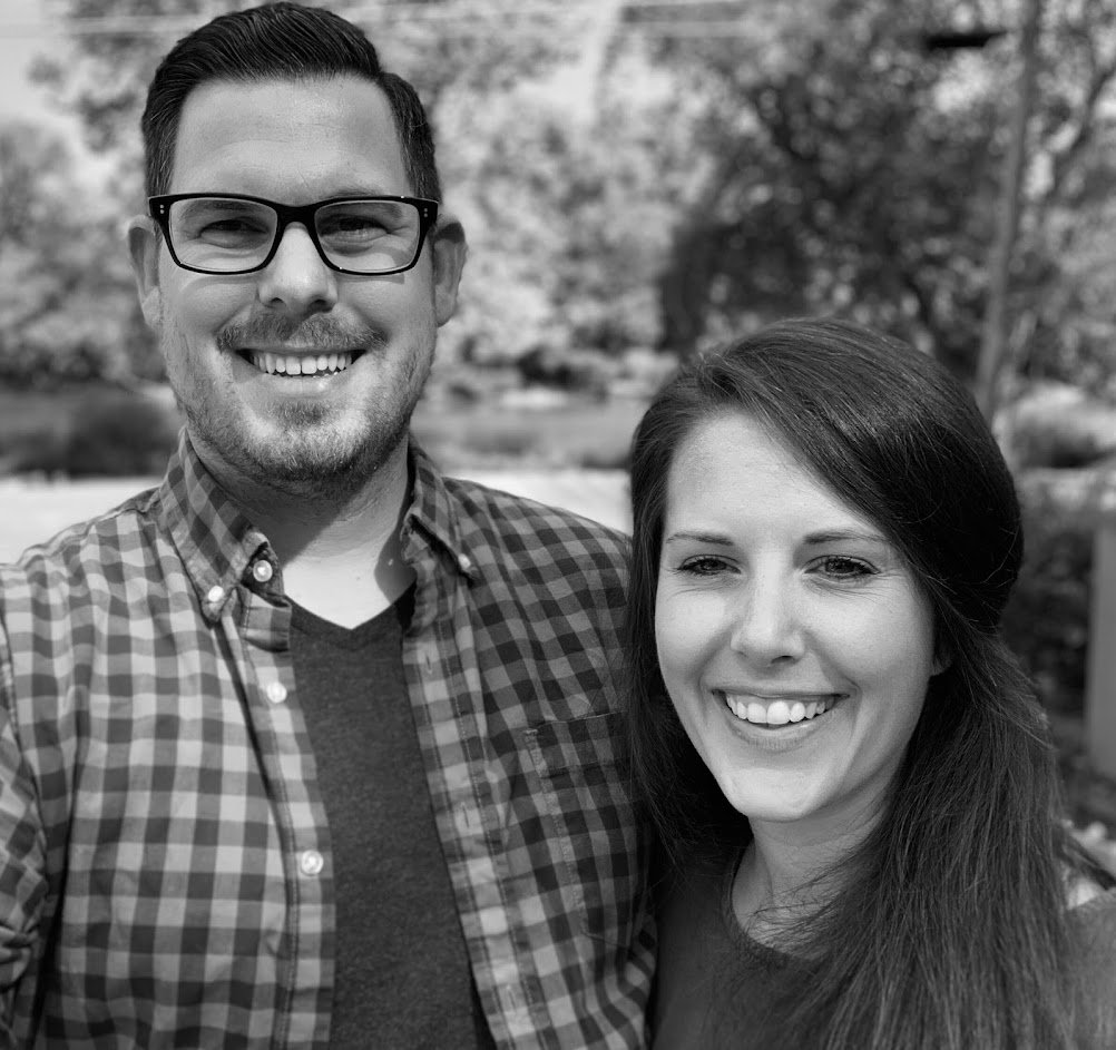 A man in glasses and a checkered shirt stands next to a woman with long hair, both smiling at the camera outdoors with trees blurred in the background, as if posing for a candid shot outside professional studios.
