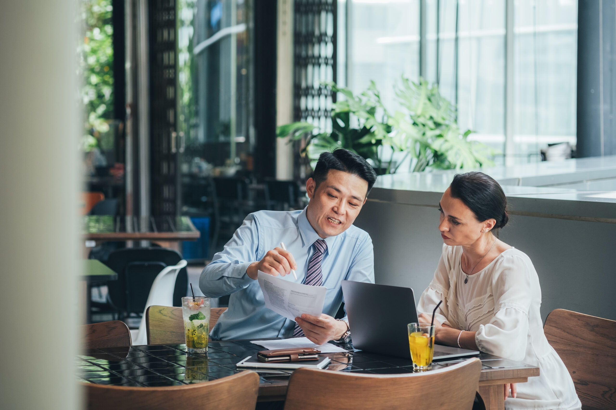 Two people sit at a table with drinks, papers, and a laptop, discussing plans for the future in a bright, modern cafe with large windows and plants in the background.