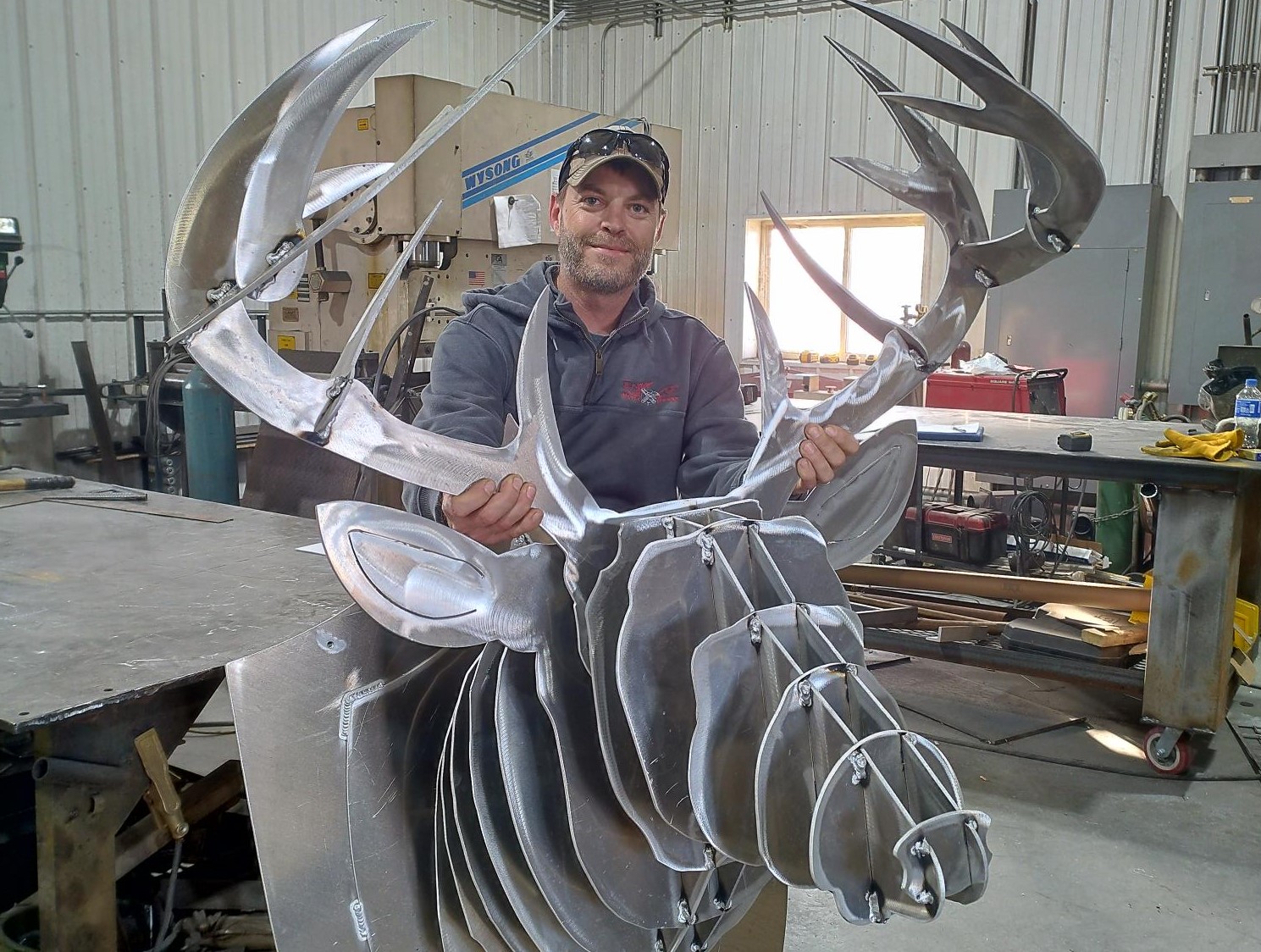 A man in a workshop holds a large, sculptural metal deer head with antlers. Crafted from layered metal sheets, the detailed piece reflects his dedication to better business practices. Industrial tools and equipment fill the background.