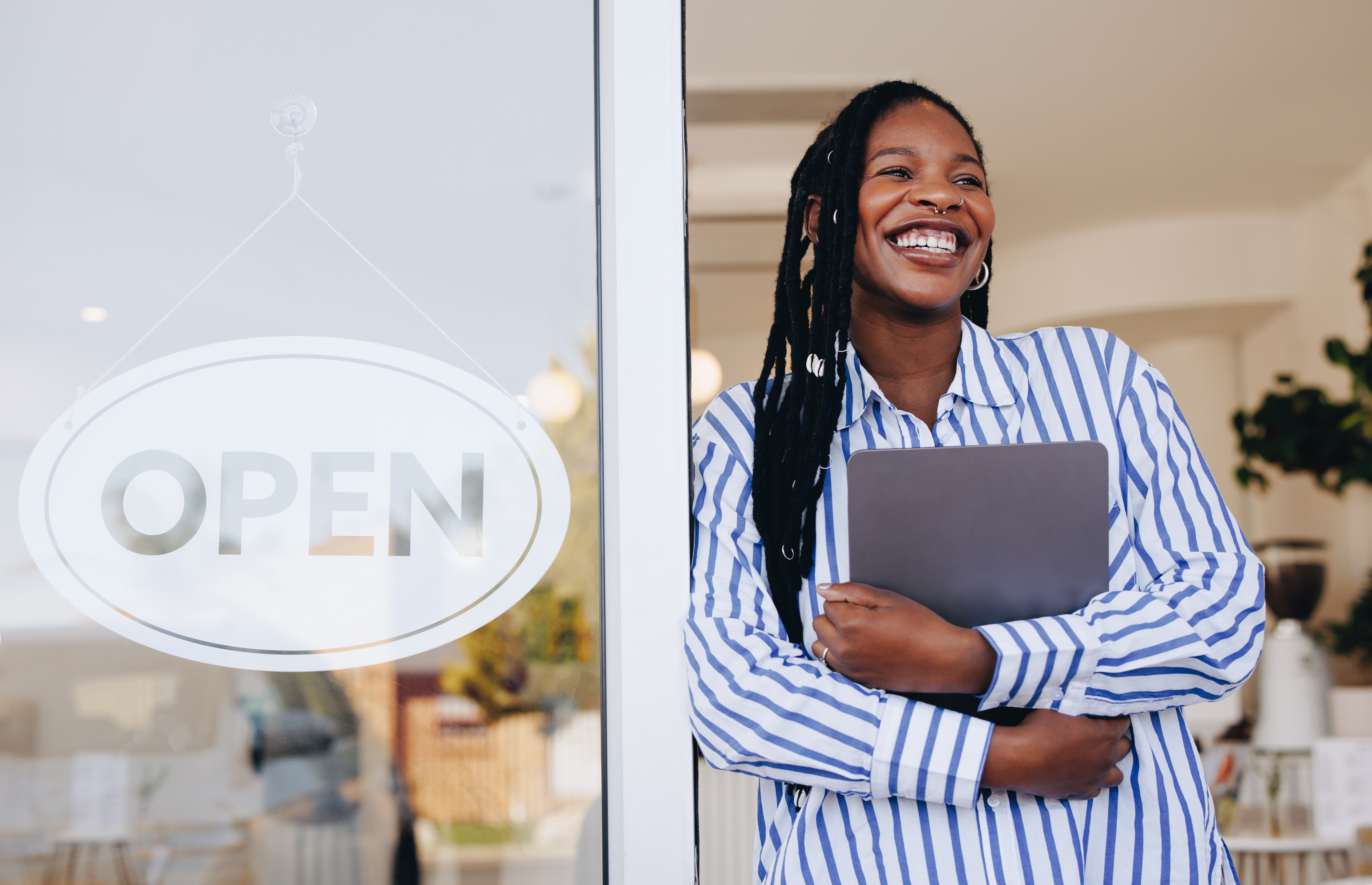 A smiling woman stands in a doorway next to an OPEN sign, holding a laptop and wearing a blue and white striped shirt, suggesting she is a tech-savvy business owner or employee.